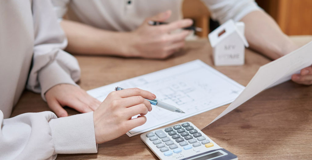 Vue en plongée d'une table avec un plan architectural d'appartement, une calculatrice et un stylo, mains d'un adulte travaillant dessus